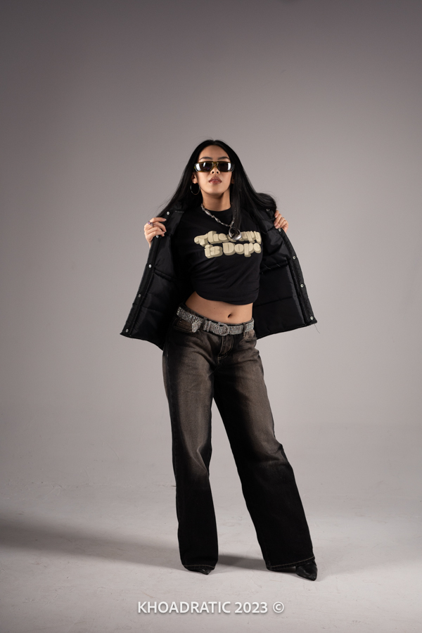 A person poses confidently, wearing sunglasses, a black jacket, and wide-legged jeans against a plain gray background.