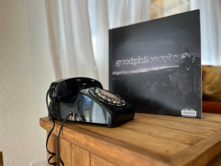 A black rotary phone sits beside a vinyl record titled "Good Philosophy" on a wooden table. White curtains are in the background.