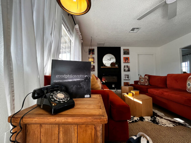A cozy living room with a red sofa, wooden coffee table, rotary phone, and a "good philosophy" album on display. Walls feature framed photos and a round mirror above the fireplace.