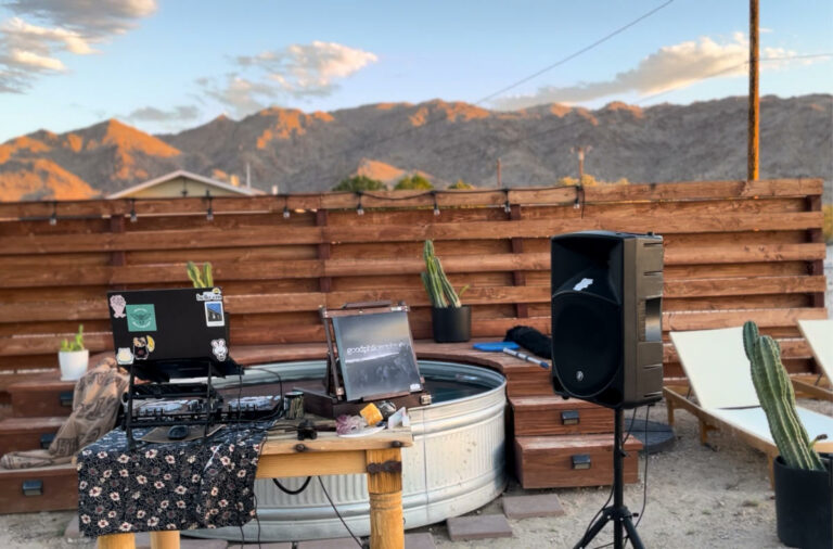 Outdoor setup with DJ equipment and speakers on a table in a desert backyard. Cacti, a metal tub, and mountains are in the background.