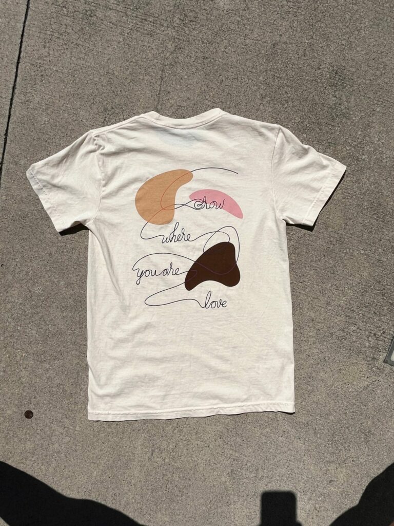 A white T-shirt with a colorful abstract design and the text "grow where you are love" on the back.