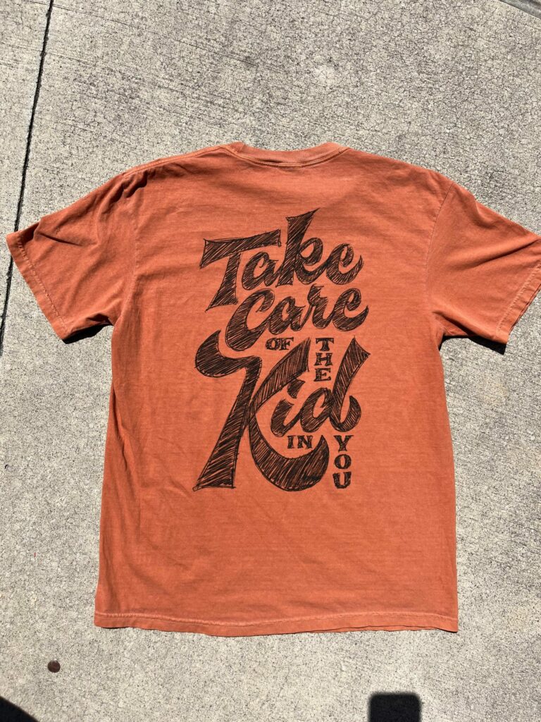 An orange T-shirt with the text "Take care of the kid in you" written in large black script on the back.