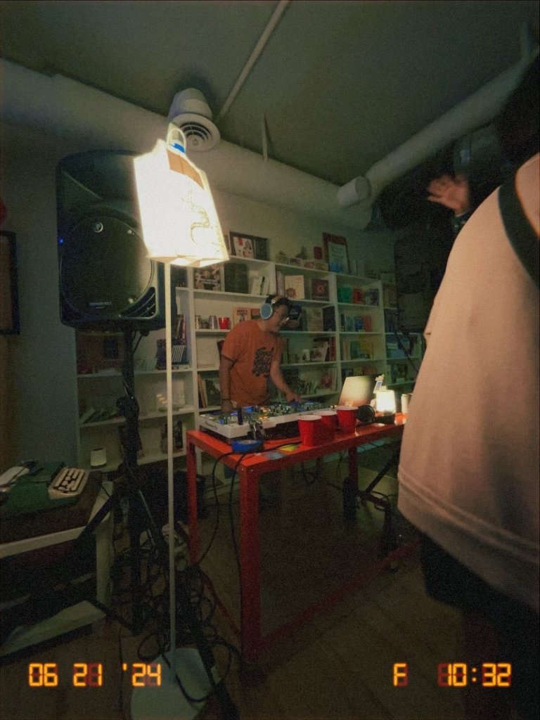 Andrew playing music on mixing equipment in a dimly lit room with shelves in the background. Timestamp in the corner reads "06 21 '24".